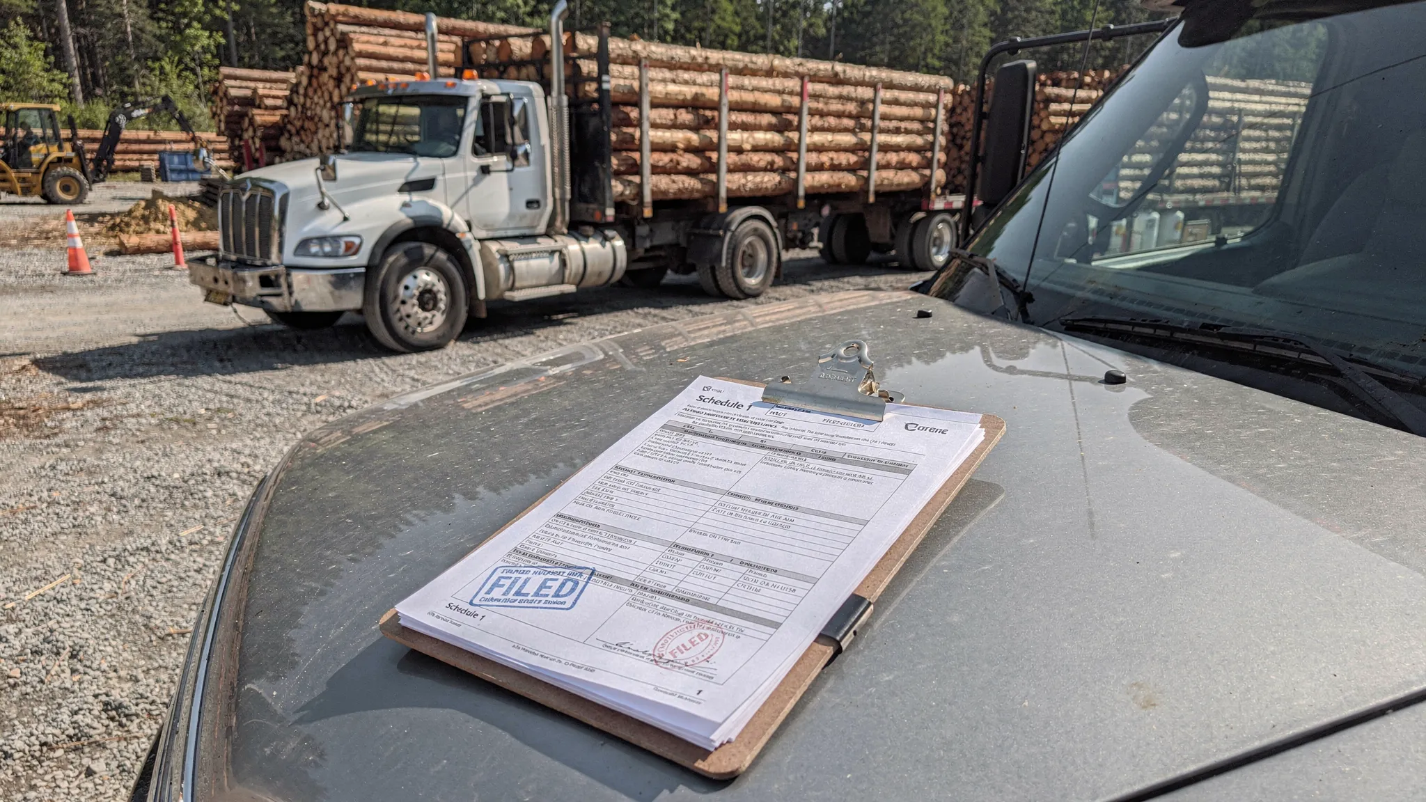 A logging truck parked near a timber yard with a clipboard and paperwork on the hood, showing a stamped Schedule 1 document as proof of HVUT filing, with stacked logs and forestry equipment in the background.